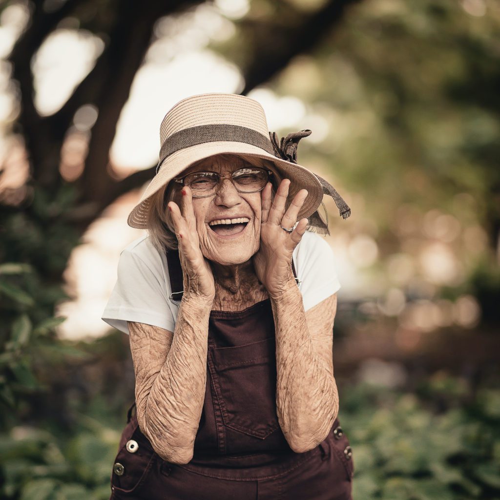An old woman with a bunch of wrinkles wearing a straw hat. Her hands are on the sides of her face and she looks like she is yelling at some. Smiling real big | who does god say I am | Positively Jane