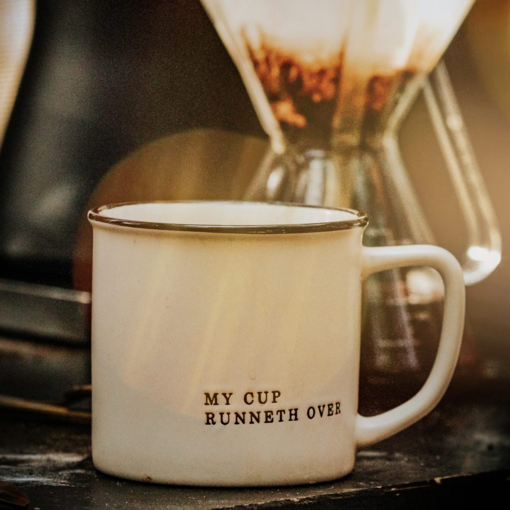 white coffee mug next to a chemex coffee maker. the mug says 'my cup runneth over' on it | gratitude changes attitude | Positively Jane