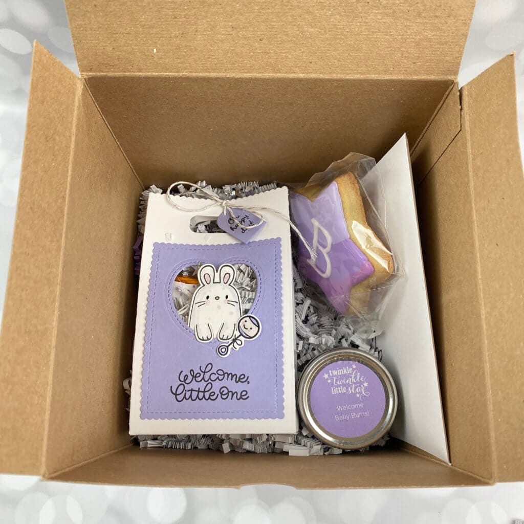 Box filled with baby shower treats. Little bag, cookie, candle and an envelope with white paper shred on the bottom | hosting a virtual baby shower | Positively Jane 
