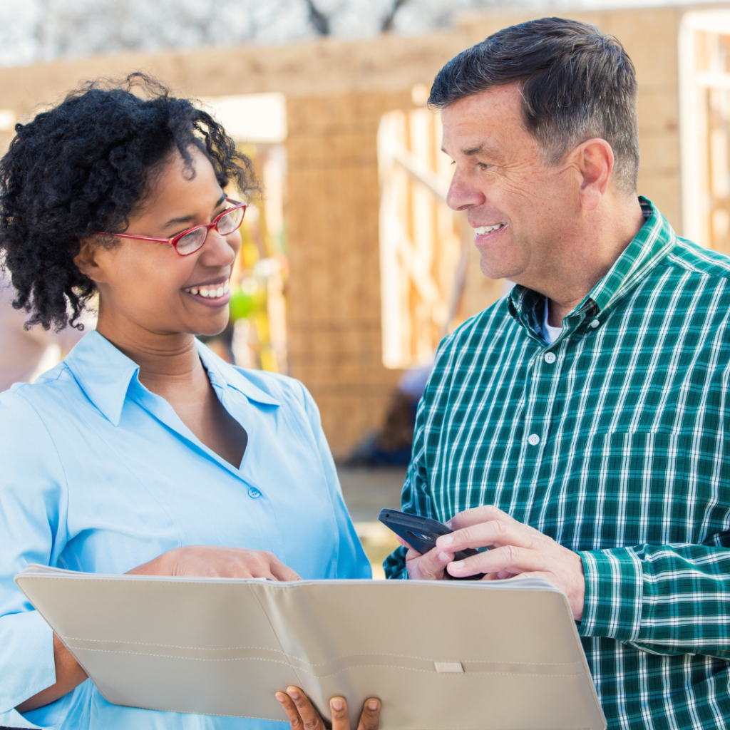 a man and a woman looking at house plans with a house in framing stages behind them | choosing a home builder 